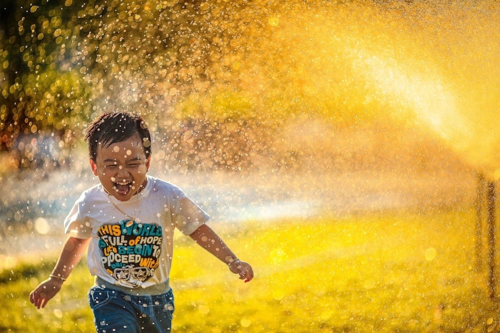 Happy kid running through a water sprinkler during a hot summer day