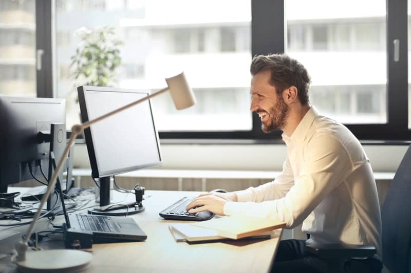 man-in-white-dress-shirt-sitting-on-black-rolling-chair-while-facing-black-computer-set-and-smiling