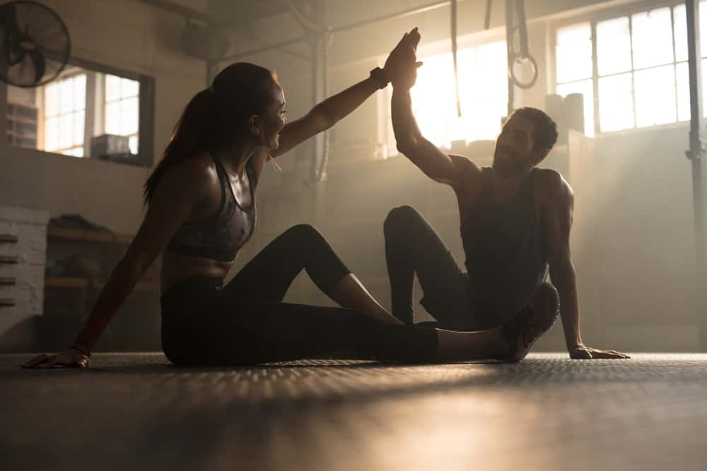 A woman and a man are sitting on the floor of a gym and giving each other high five.