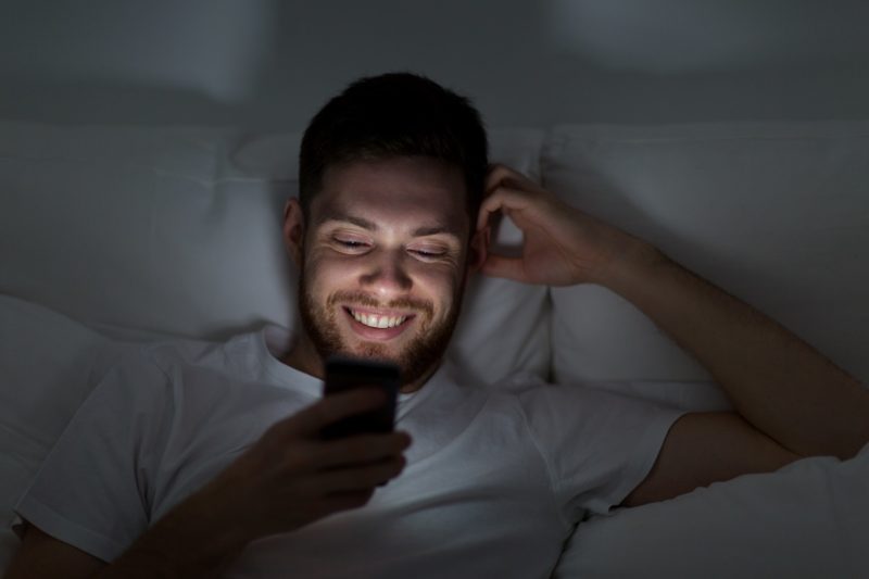 happy young man with smartphone in bed at night