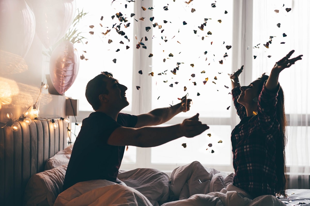 Cheerful young man and woman throw shiny confetti celebrating anniversary in light hotel room