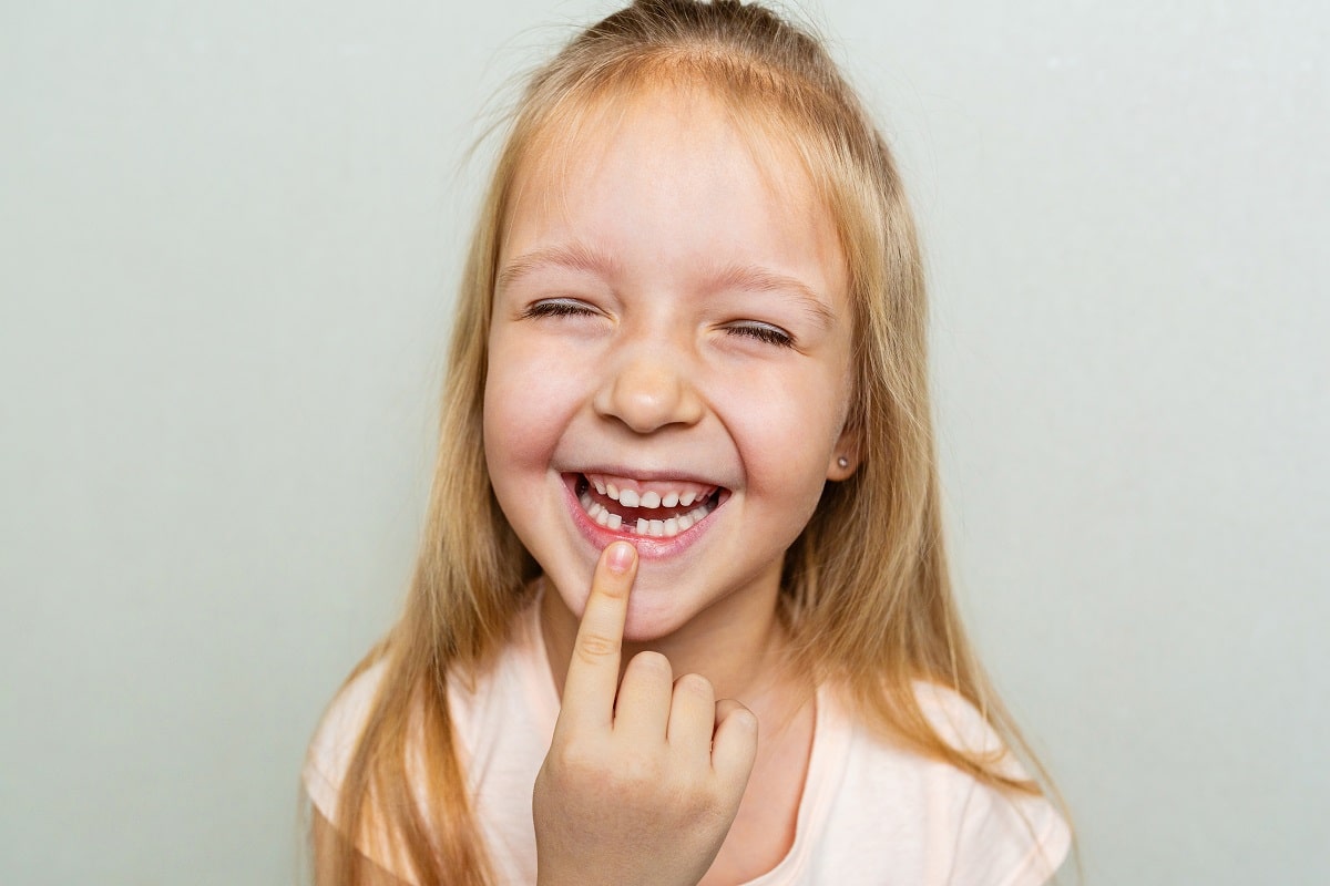 Happy little Caucasian girl with blonde hair showing missed milk tooth on gray background