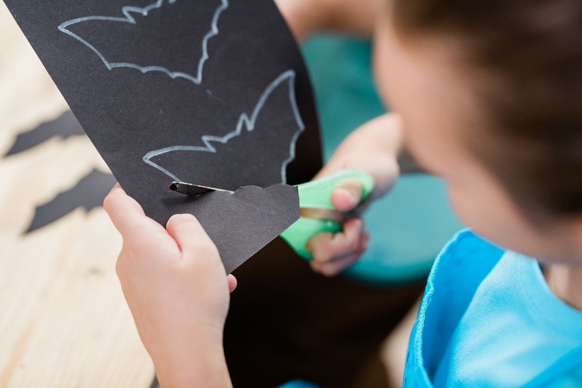 Hands of elementary schoolgirl with scissors cutting out bat from black paper